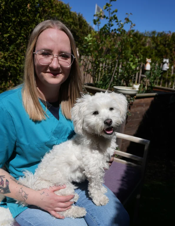 Ein Porträtfoto von Denise Uster. Sie sitzt in einem Garten und hält die kleine, weiße, lockige Hündin Mia auf dem Schoß. Sie trägt eine Brille und eine türkisfarbene medizinische Tunika über einem schwarzen Shirt. Mia sitzt aufrecht und schaut nach vorne. Der Hintergrund ist ein sonniger Garten mit Hecke, Zaun und Gartenfiguren.