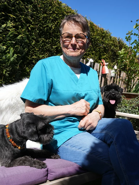 Petra Irmscher sitzt entspannt in einem Garten, flankiert von zwei schwarzen Schnauzer-Hunden, Balou und Buffy. Sie trägt eine türkisfarbene medizinische Tunika über einem hellen Shirt und Jeans. Einer der Hunde sitzt links von ihr und schaut nach rechts, der andere sitzt rechts von ihr und schaut nach vorne mit herabhängender Zunge. Der Hintergrund zeigt eine Hecke, einen Holzzaun mit Gartenfiguren und einen blauen Himmel.